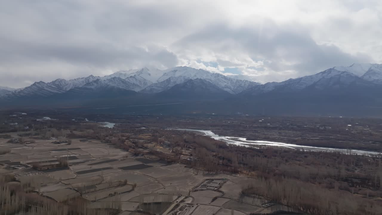 Den frosne Indus-flod snor sig gennem Leh, en højtliggende dal i Ladakh, Indien, med landbrugsmarker og spredte bebyggelser. Langsom indzoom mod sneklædte Himalaya-toppe og overskyet vinterhimmel.