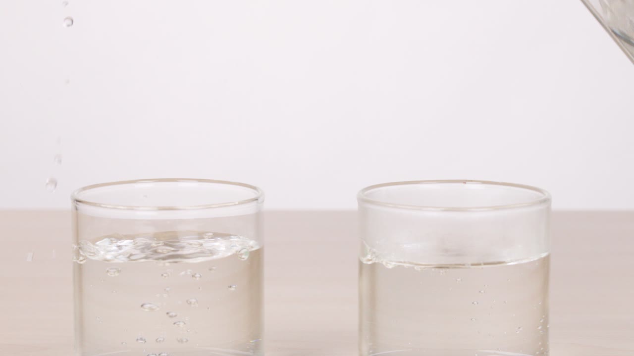 Water is poured from a glass pitcher into two empty glasses on a light surface