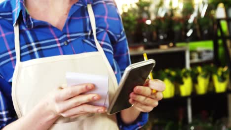 female florist writing an order on notepad while using mobile phone