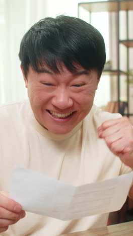 asian man smiling while reading open letter on paper glad about good news joyful reaction at desk