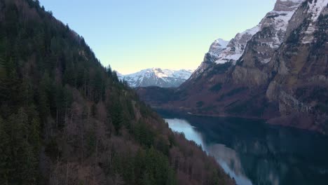 Aerial-slow-flying-over-a-lake-and-forest-with-snowy-background-mountains-in-the-Alps,-Switzerland