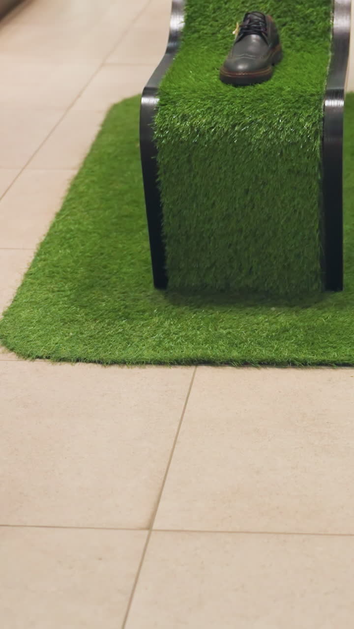 un zapato negro y marrón colocado en un soporte único cubierto de hierba artificial verde. la pantalla es parte de una configuración de una tienda minorista con ropa visible en el fondo y una persona caminando por delante