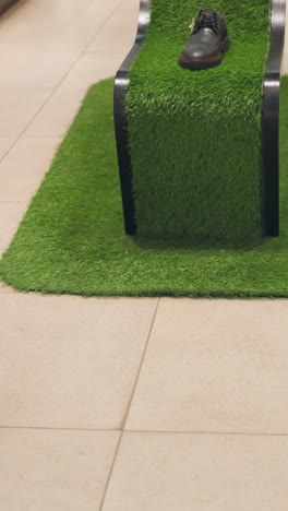 un zapato negro y marrón colocado en un soporte único cubierto de hierba artificial verde. la pantalla es parte de una configuración de una tienda minorista con ropa visible en el fondo y una persona caminando por delante