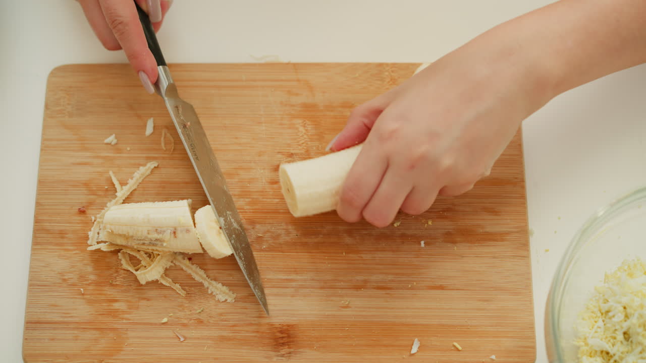 Hand view of chef carefully cutting spoiled part of banana on cutting board and slicing remaining portion into tiny round shapes, preparing fresh ingredients in bright, organized kitchen environment