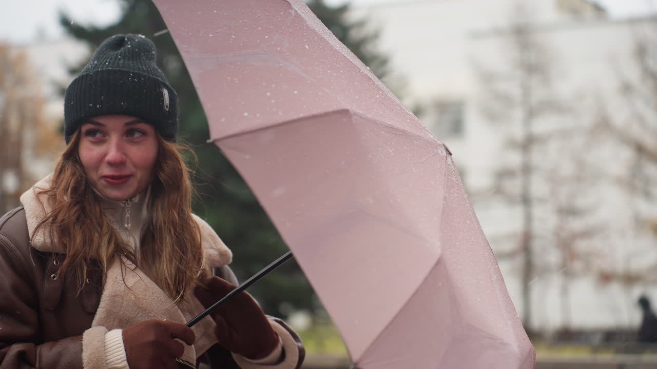 Female wearing black knit cap and brown shearling jacket holding umbrella as wind blows gently with light snowfall, strands of hair in motion, capturing quiet winter moment with calm serene mood outdoors
