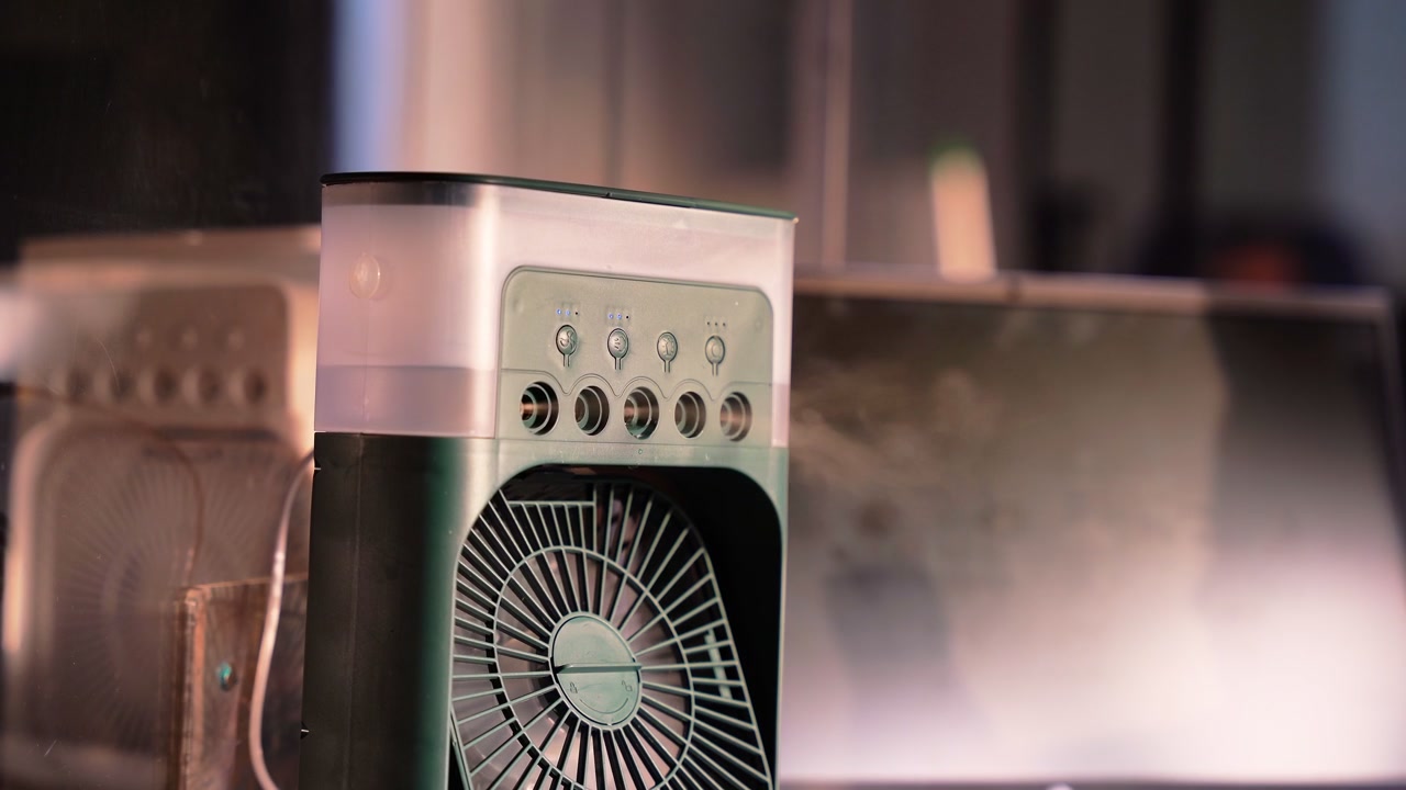 A portable green evaporative air cooler sprays water mist from five nozzles in a dim modern office interior at night near a computer monitor. Cinematic shallow depth of field.