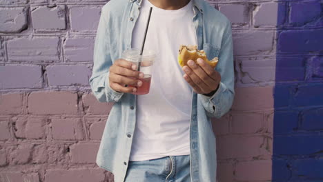 市内のカラフルなレンガの壁にもたれて、おいしいハンバーガーを食べ、プラスチック製のカップに冷たい飲み物を持っている幸せな若い男 1