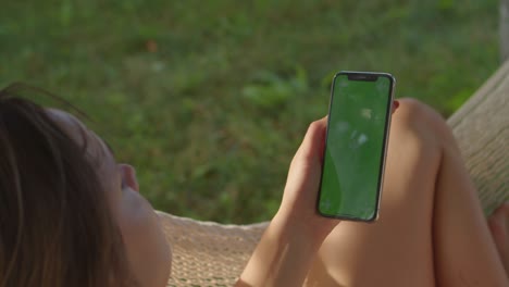 young women rests in hammock looking into vertical phone with green screen