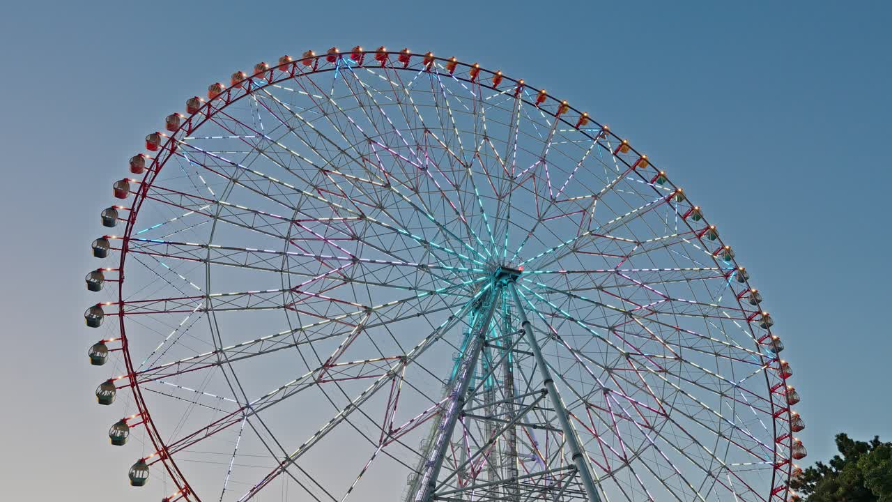 A vibrant, slightly low-angle shot of the Diamond and Flower Ferris Wheel featuring aqua and violet lighting against the bright sky