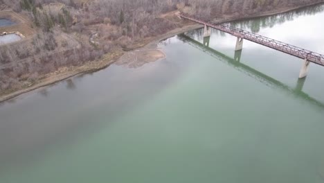 1-3-Vuelo-Aéreo-Sobre-El-Lúgubre-Parque-Fluvial-Por-Un-Puente-Multimaterial-Para-Peatones-De-Madera-De-Hormigón-Y-Acero-En-Una-Tarde-De-Otoño-Donde-Aparecen-Aguas-Contaminadas-Merky-Cúmulos-De-Desechos-De-Baño-Derramados-De-Aceite