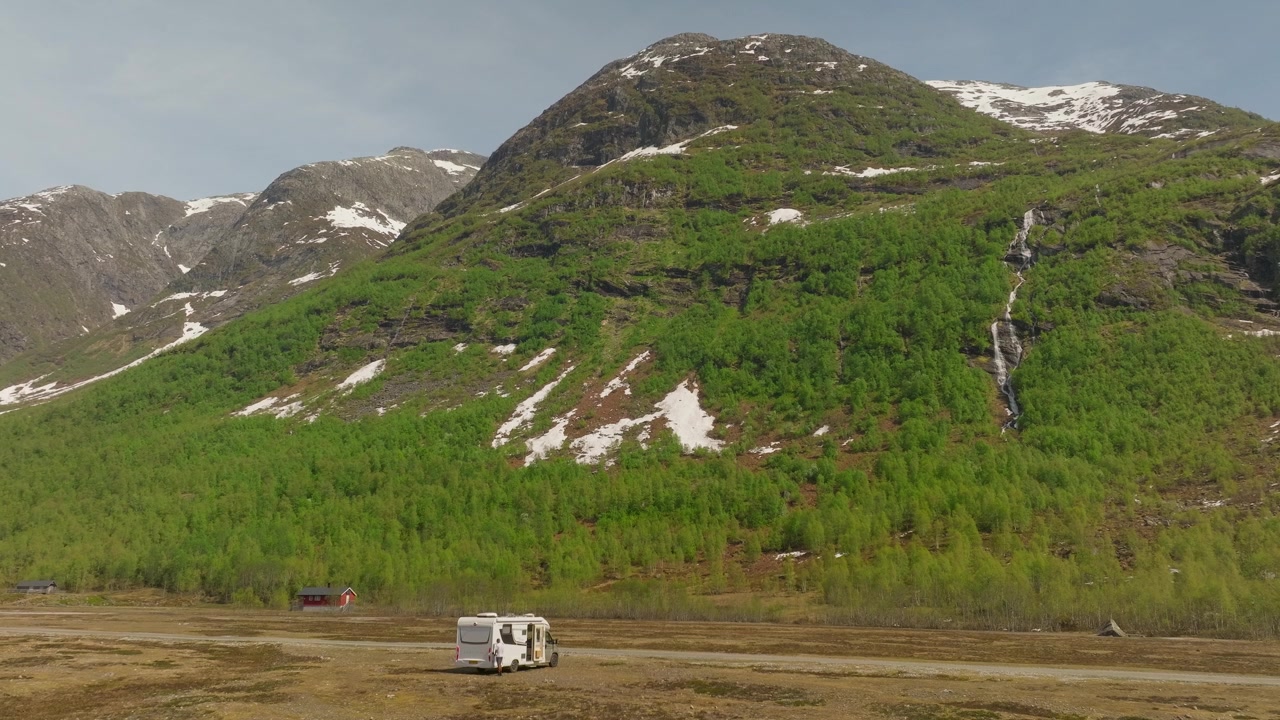 Un viaggiatore scende da un camper parcheggiato vicino a una cascata di montagna, scaricando l'attrezzatura mentre è circondato da un paesaggio alpino mozzafiato e cieli blu in Norvegia.