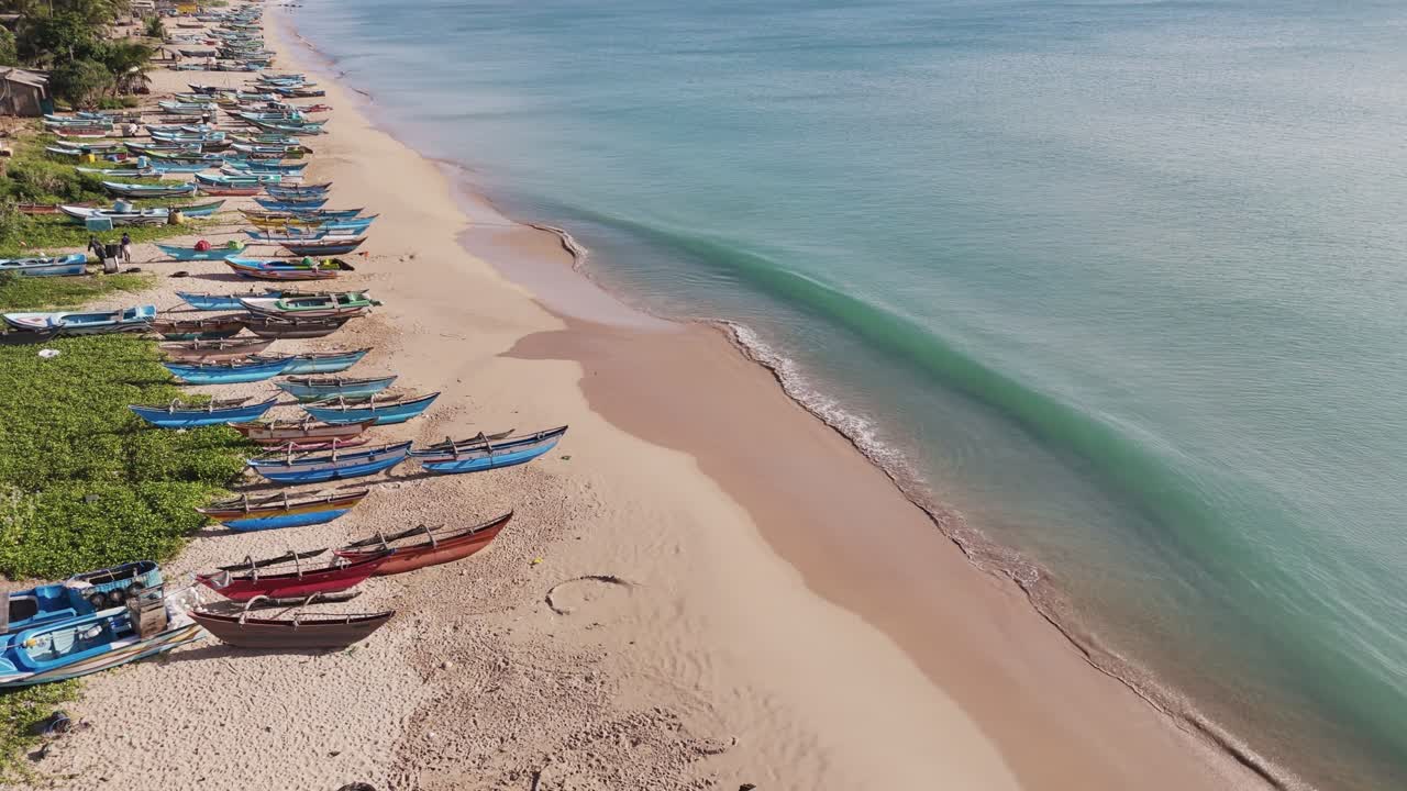 Green blue wave formations lapping the white sandy shoreline with fishermen’s boars onTrincomalee beach, Sri Lanka. Aerial static .