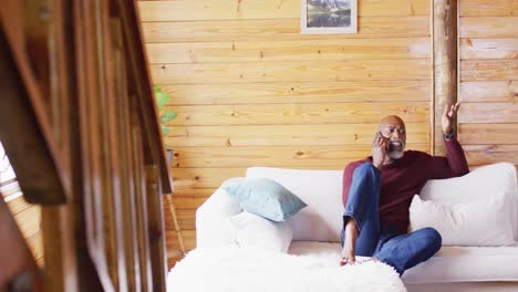 Happy-senior-african-american-man-spending-time-in-log-cabin,-sitting-on-sofa-and-using-smartphone