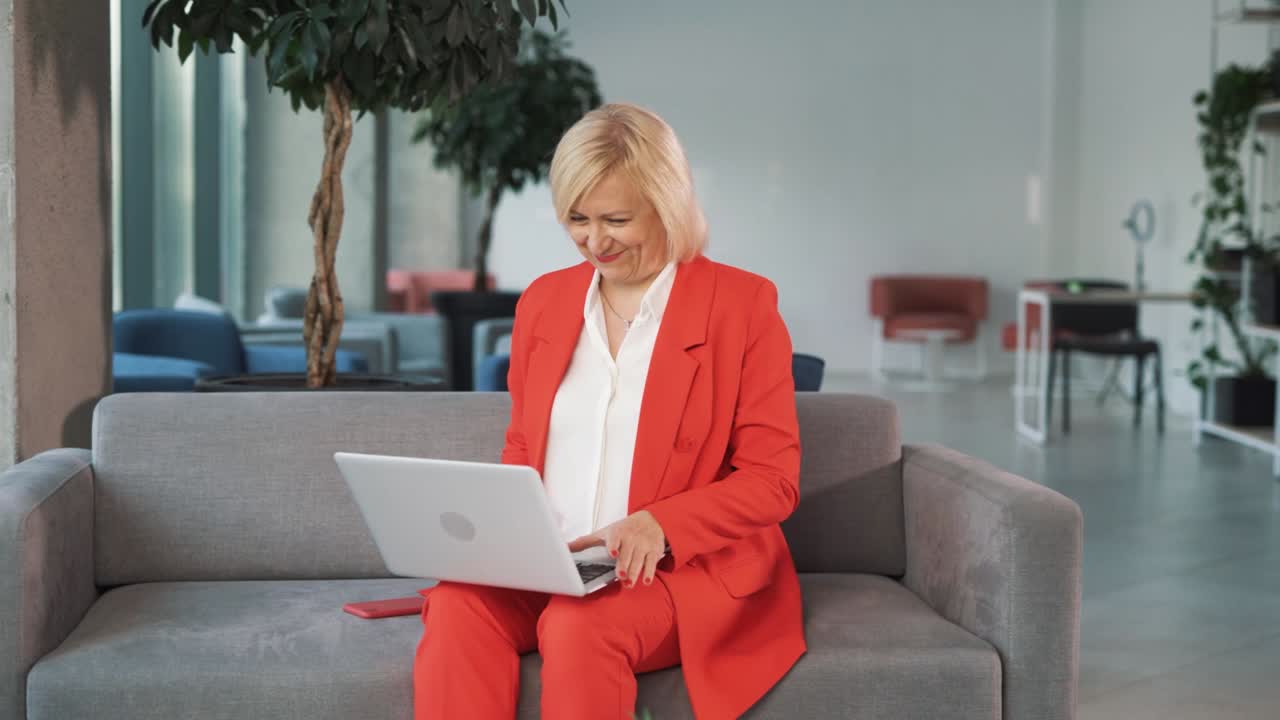 older woman in a red business suit, sitting on a couch in a business center, turns on her laptop and begins a video call, greeting with hello