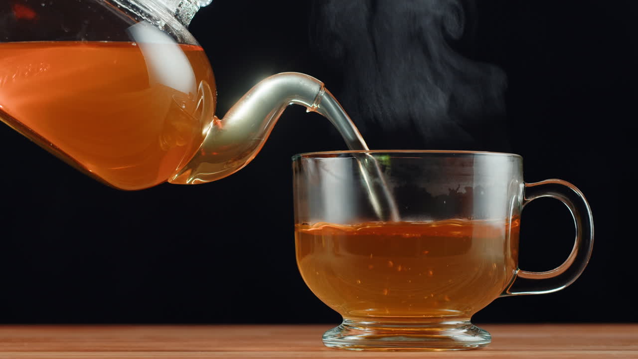 Hot tea being poured from a glass teapot into a clear glass cup, with steam rising
