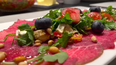 close up tilt down shot of beautiful served carpaccio meat with arugula, tomatoes, blueberries and roasted seeds