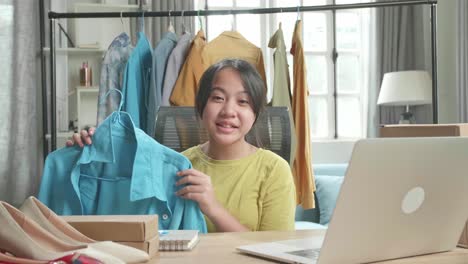 joven asiática vendedora en línea recomendando una camisa mientras usa la computadora para vender ropa en casa