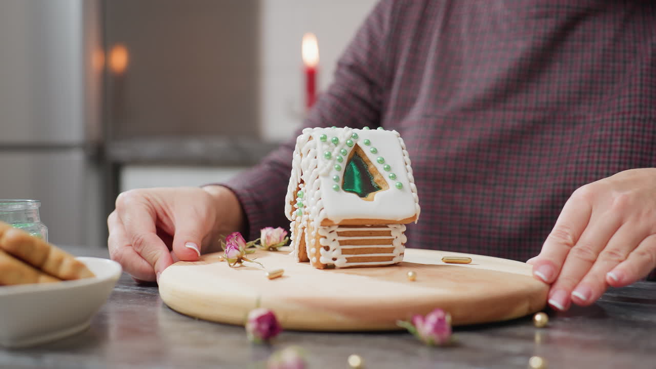 caterer rotando cuidadosamente y mostrando una casa de pan de jengibre bellamente decorada con hielo blanco y cuentas verdes, bandeja de madera, salpicaduras doradas, flores secas y luz de vela borrosa en el fondo