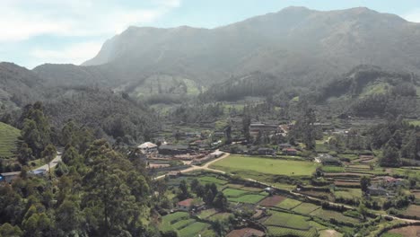 Rural-town-and-farm-fields-amidst-high-mountainous-landscape-and-tea-garden-fields-in-Munnar,-India---Aerial-Orbit-shot