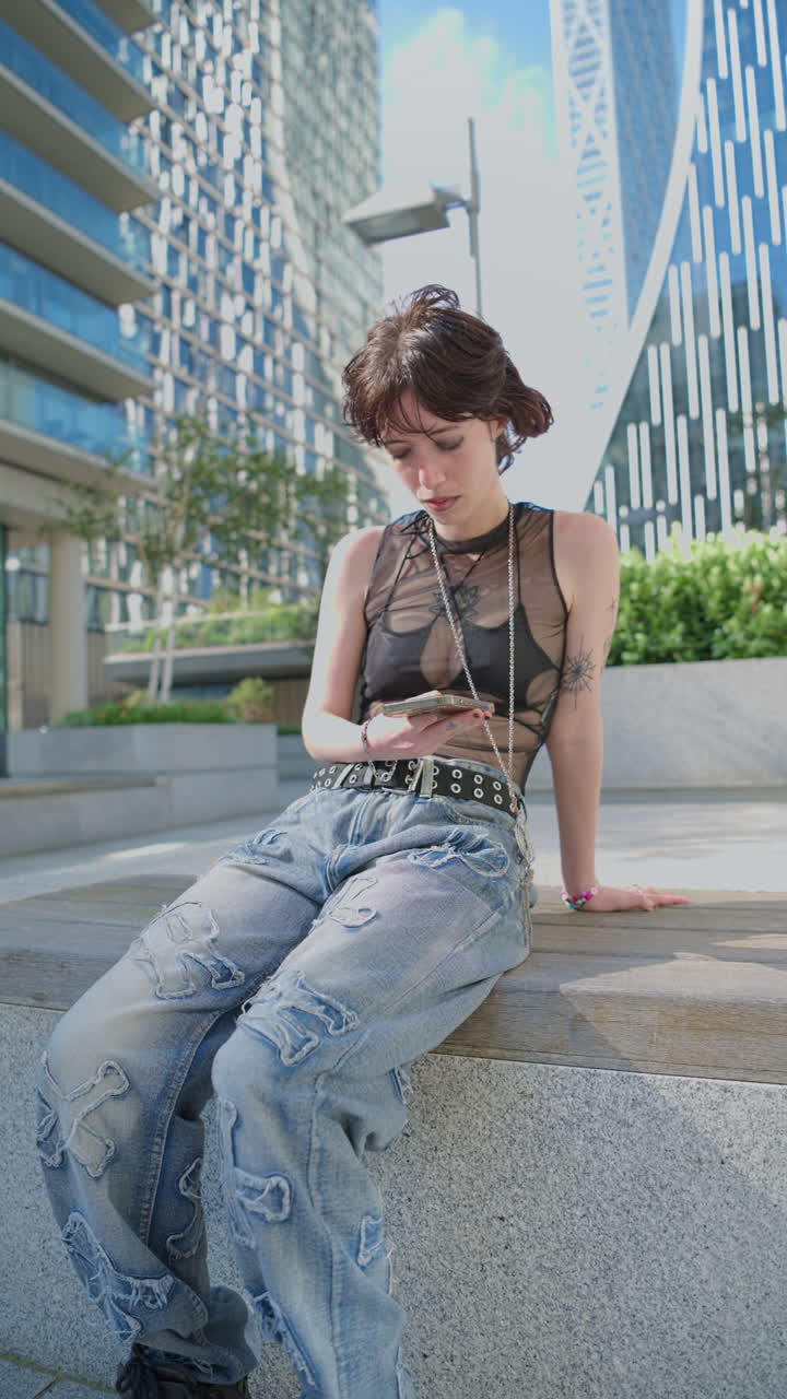 retrato vertical de moda al aire libre de una mujer joven de estilo alternativo con tatuajes sentada en un banco enviando mensajes por teléfono móvil en canary wharf, londres