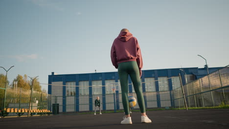 vista trasera de una mujer rebotando el voleibol con dos mujeres mirando en el fondo, el edificio visible detrás de ellos en la cancha de voleibol al aire libre, mostrando una sesión de práctica