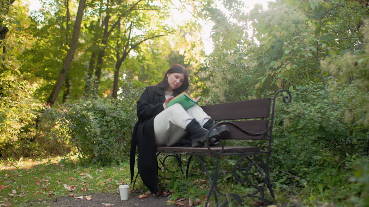 Zoom in on elegant woman on park bench with legs up reading book, pausing to adjust hair; paper cup on ground beside, surrounded by green foliage and fallen leaves, warm sunlight, cozy coat and boots