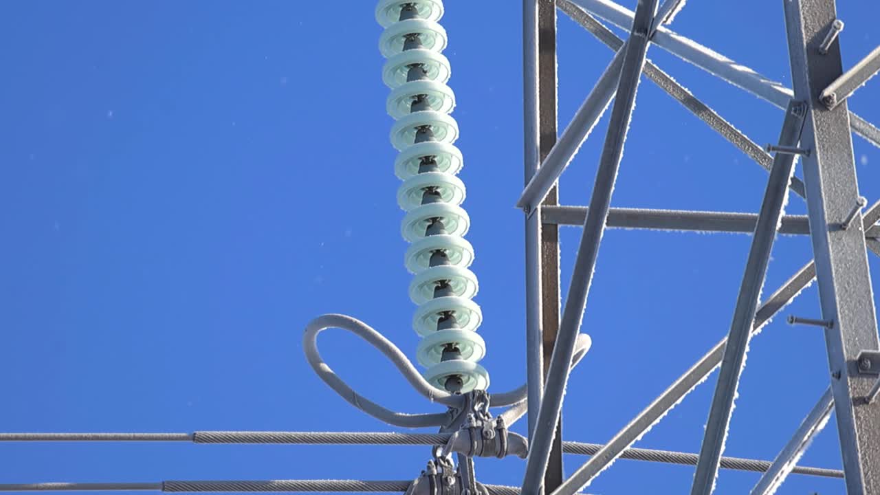 Close view of high voltage suspension insulators coated in ice and hoarfrost on a metal power pylon, sparkling in bright winter sunlight against a deep blue sky