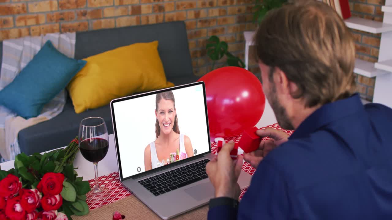 Casal caucasiano em videochamada de Dia dos Namorados, homem mostrando anel para mulher sorrindo na tela do laptop.