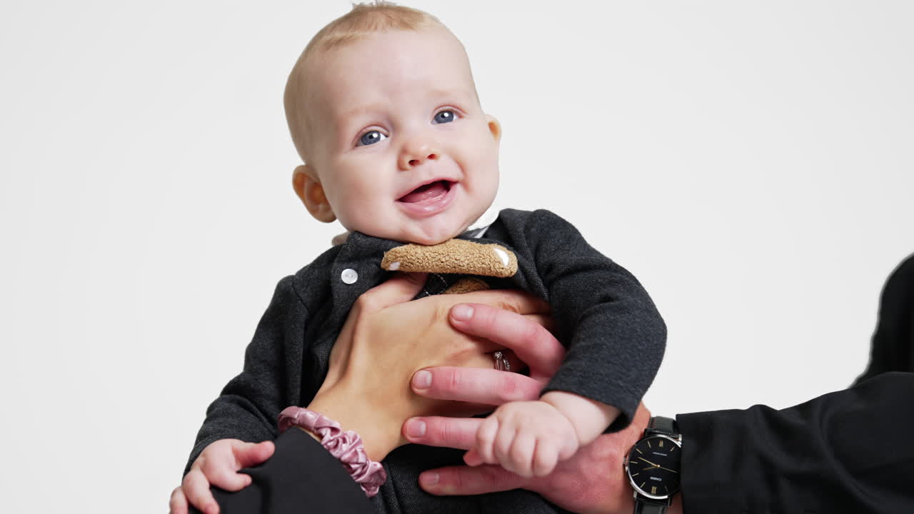 Male and female hands hold beautiful smiling Caucasian infant. Mother and father kiss their lovely son on the head. White backdrop.