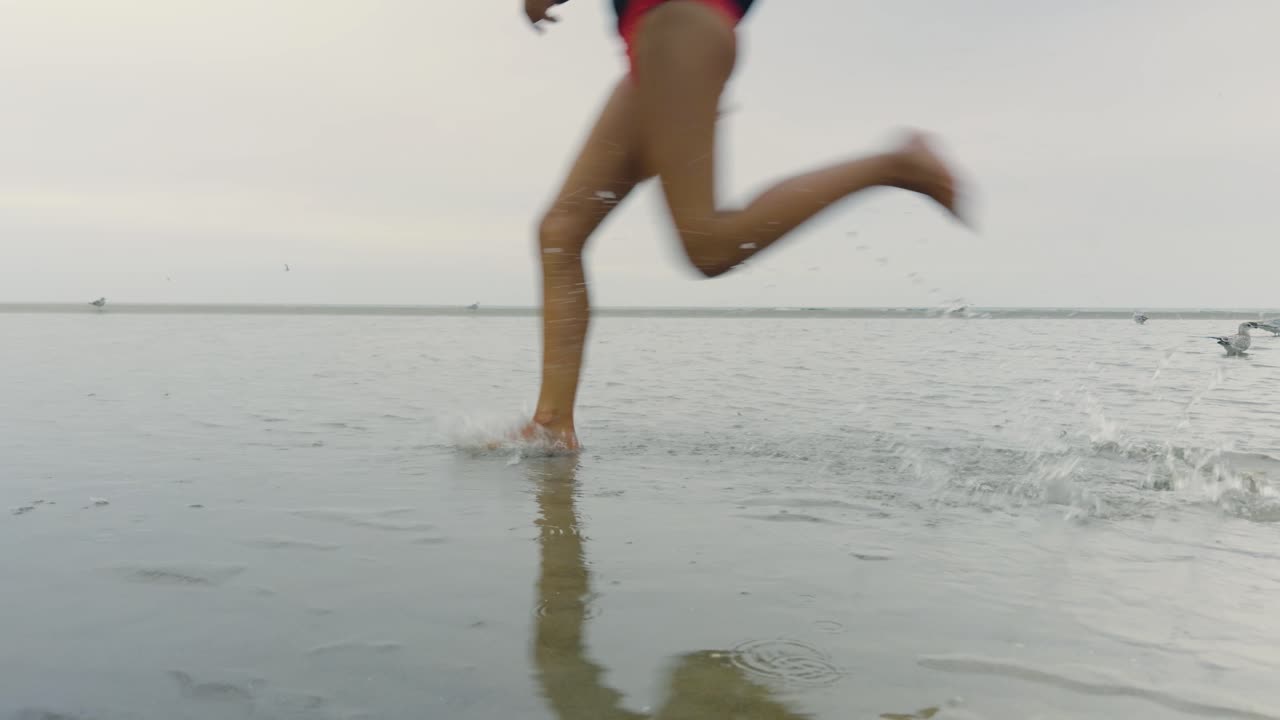 Young girl runs through shallow water at low tide, splashing under an overcast foggy sky, with seagulls wading along the beach shoreline