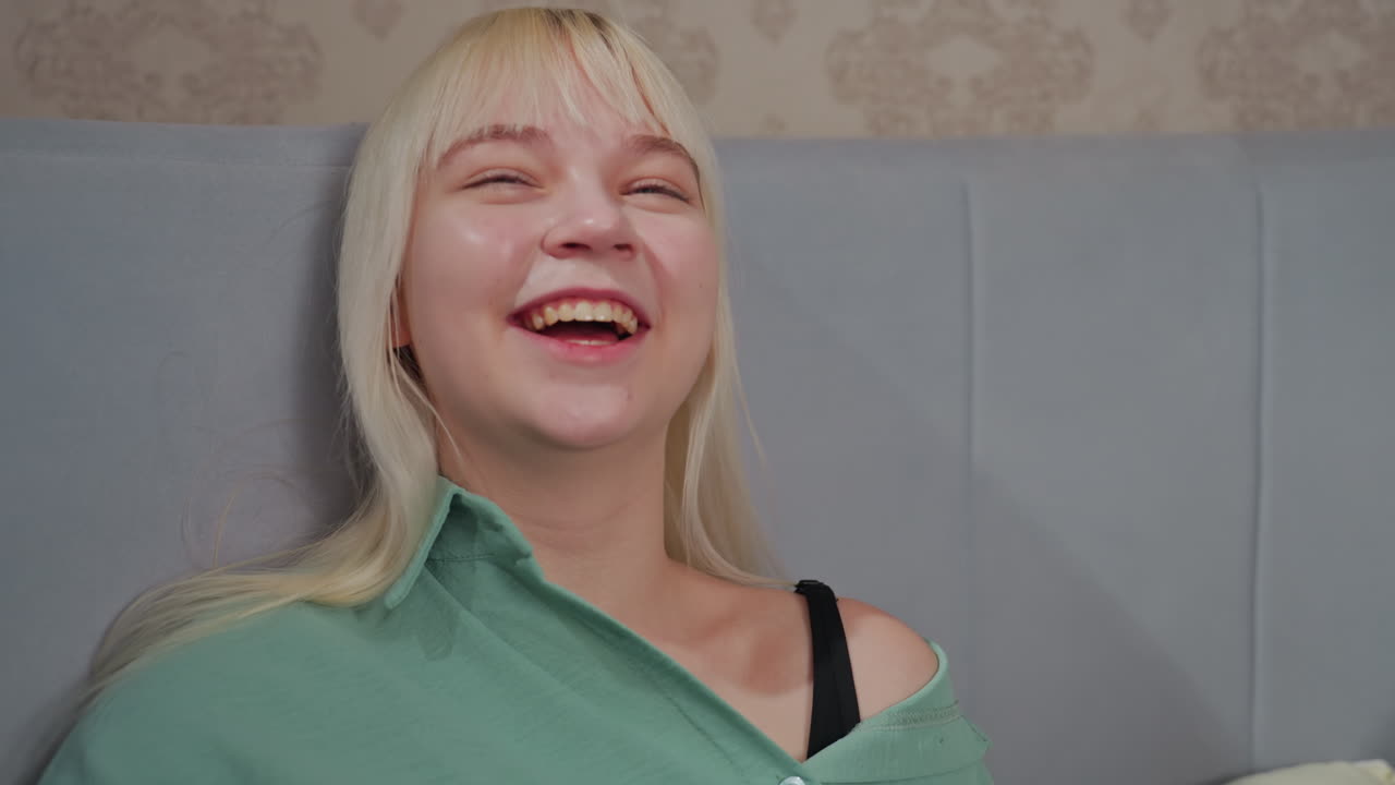 Two college girls laughing joyfully while lying on bed in relaxed setting, one holds bowl of popcorn, both enjoying lighthearted moment together with soft lighting and cozy pillows in casual bedroom