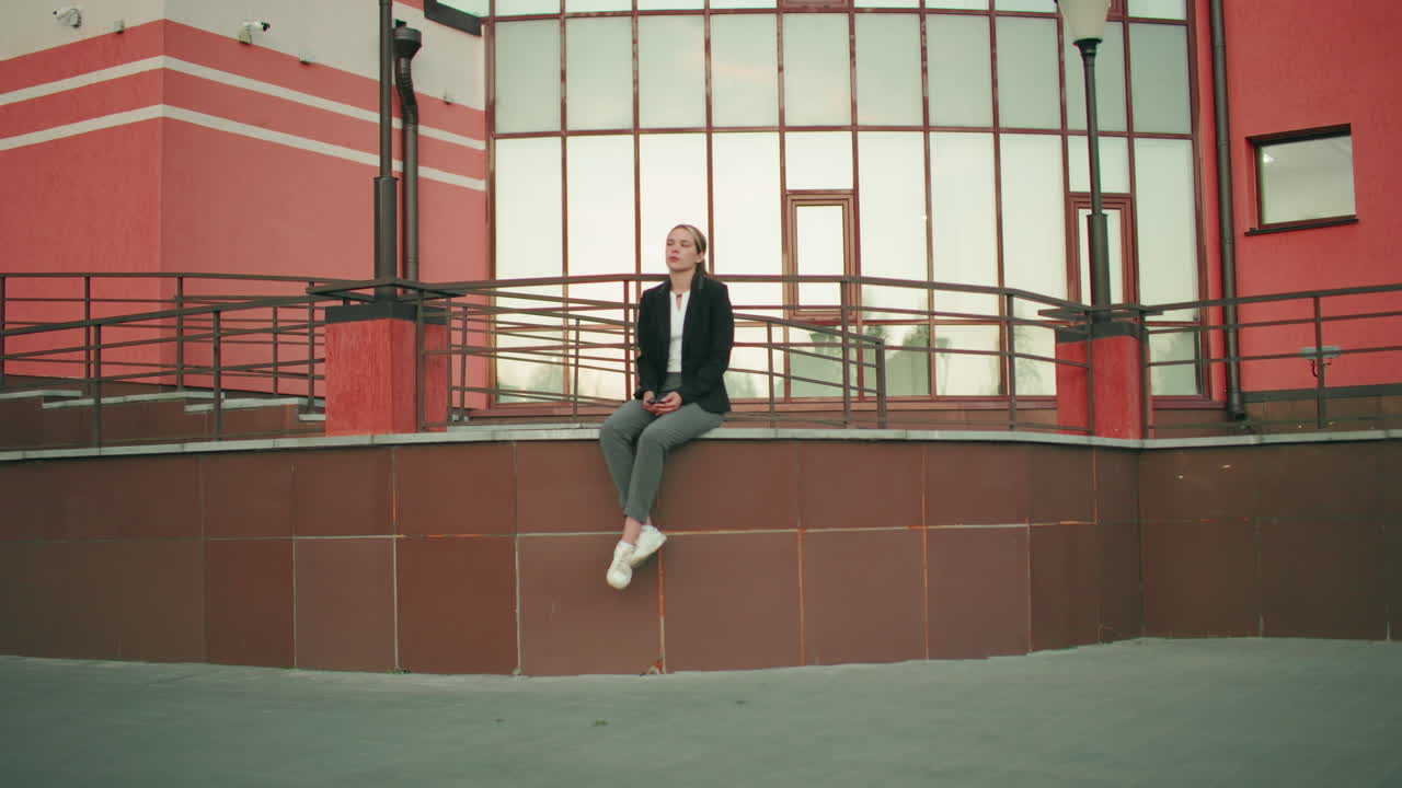 Woman seated alone on fence in solitude with legs crossed at ankle, looking at phone in hand, appearing thoughtful in front of modern building with lamp posts and railings on tiled platform