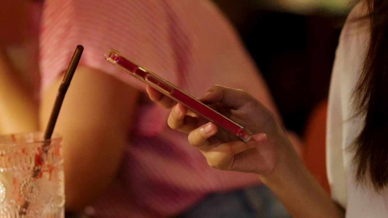 Una giovane donna interagisce con il suo smartphone accanto a una bevanda a un tavolo di ristorante illuminato caldamente, con un'altra persona in abbigliamento casual nelle vicinanze.