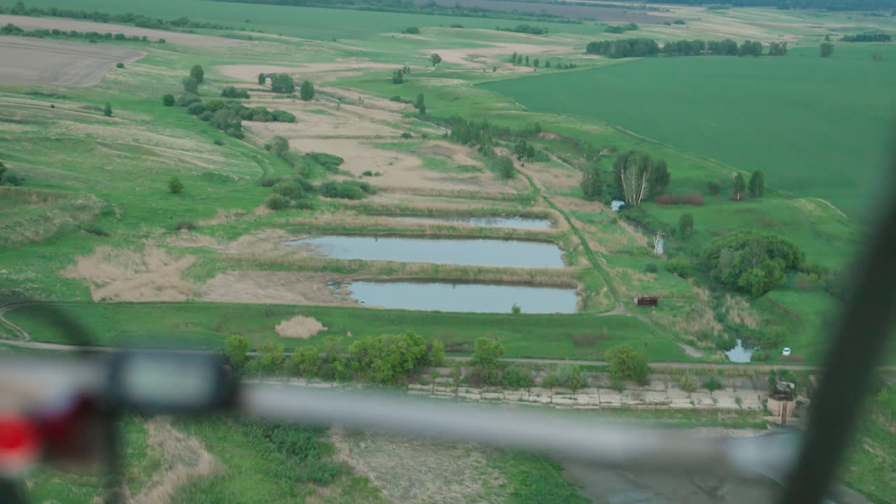 overhead drone shot capturing person holding handheld camera filming expansive green rural fields dotted with ponds and winding dirt paths under soft daylight