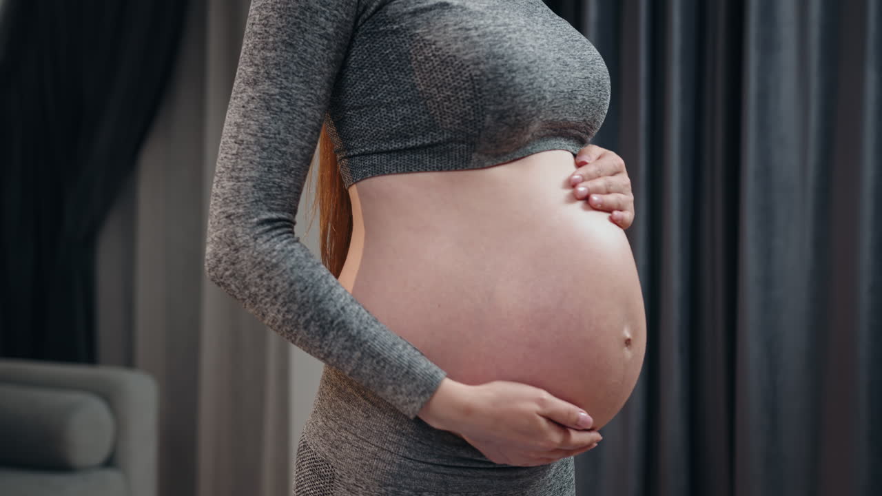 Pregnant woman in grey athletic wear cradles her rounded belly with both hands indoors with soft