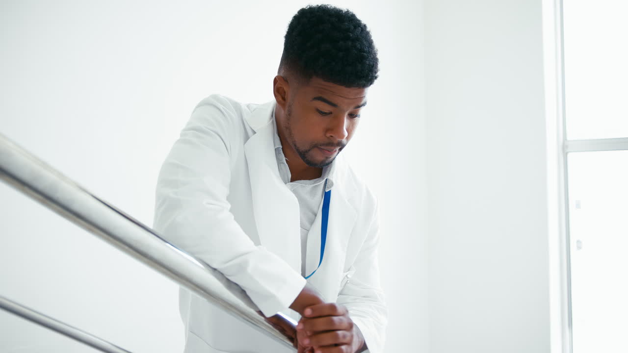 Medico maschio stressato in camice bianco in piedi sulle scale in ospedale, affetto da burnout.