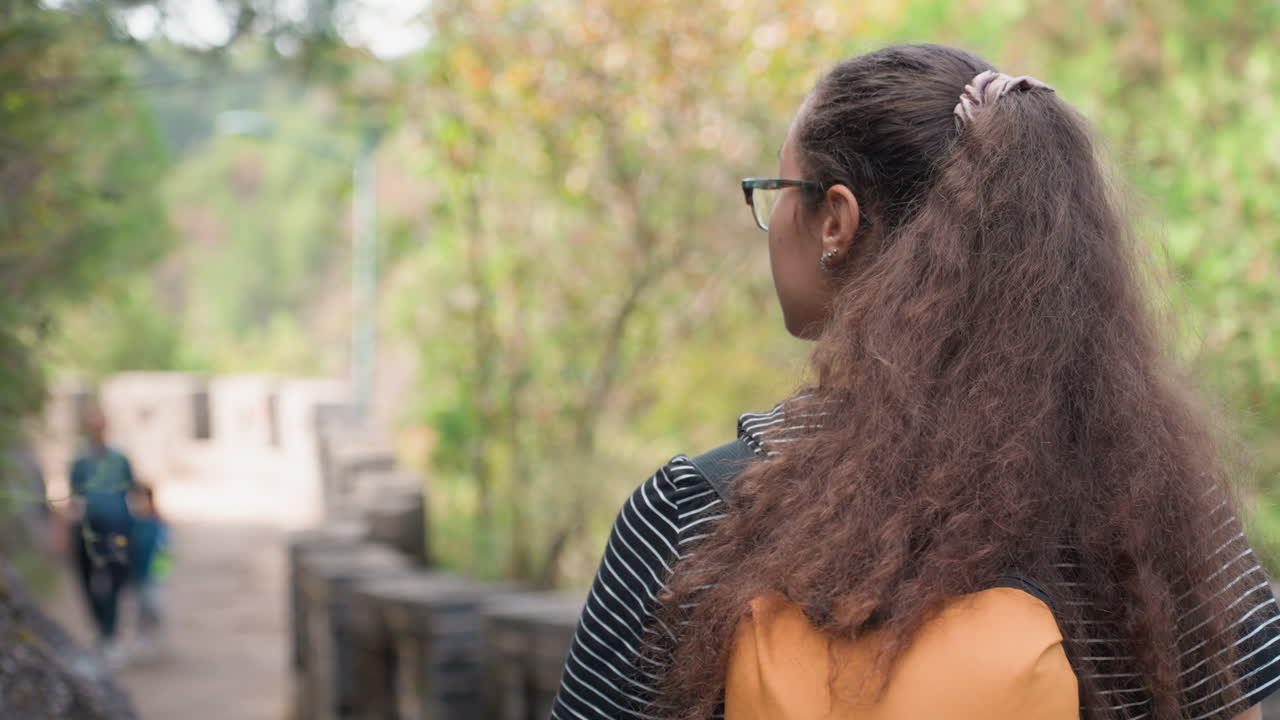 Youthful thoughtful observer. Young woman examines nature surroundings. African American lady with vibrant attire explores outdoors. Curly haired young woman with orange pack surveys mossy wall scene