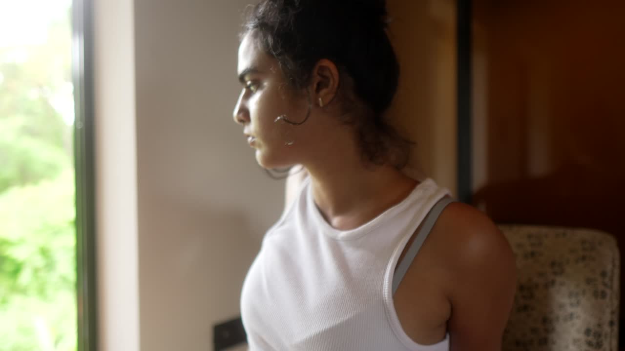 A woman in a white tank top looks thoughtfully out the window during the day