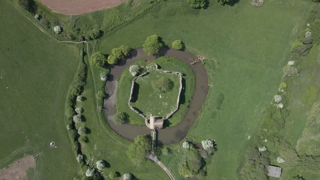 Aerial view of Stogursey Castle, a medieval castle in Somerset, England. Most of the site is in ruins, with a thatched gatehouse used for holiday's. Drone moving upwards over the ruins and moat. 4K
