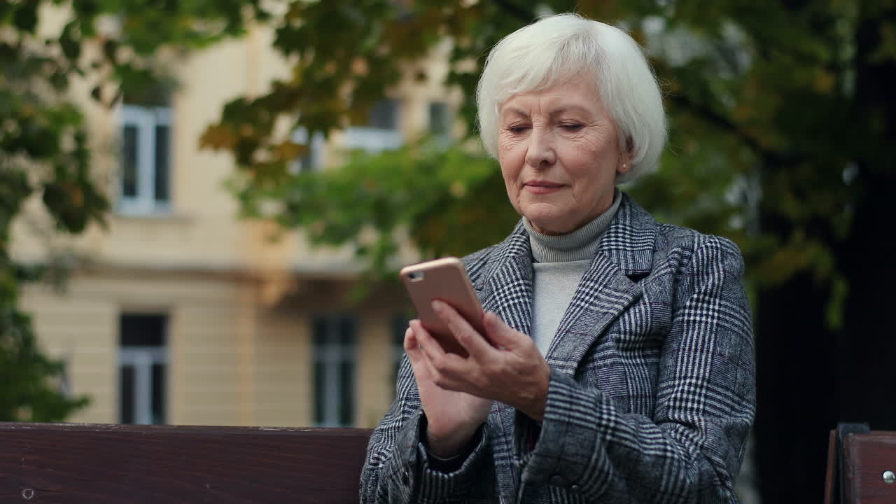 Senior Woman Sitting On The Bench In The Park And Texting While Tapping On The Smartphone