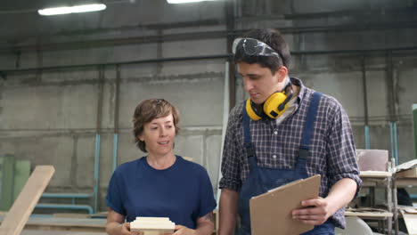 Hombre-Y-Mujer-Parados-Juntos-En-Un-Taller-De-Carpintería-Y-Discutiendo-Un-Nuevo-Proyecto-Mientras-Su-Colega-Lleva-Tablones-De-Madera-En-El-Fondo