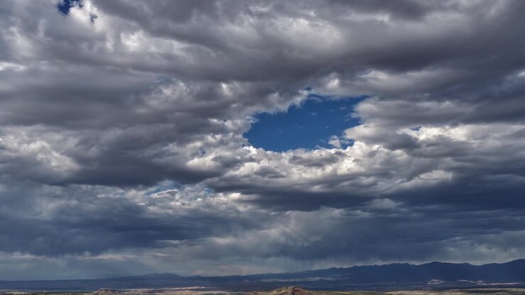 4K UHD drone timelapse of a striking hole in the sky. Heavy overcast clouds part to reveal a patch of vivid blue, shifting dynamically over a dark, vast Southwestern mountain landscape.