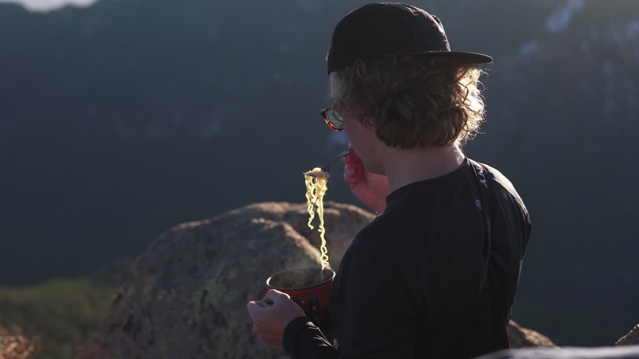 Young hiker on evening mtn summit enjoys steaming hot ramen noodles