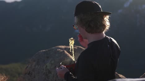 young hiker on evening mtn summit enjoys steaming hot ramen noodles