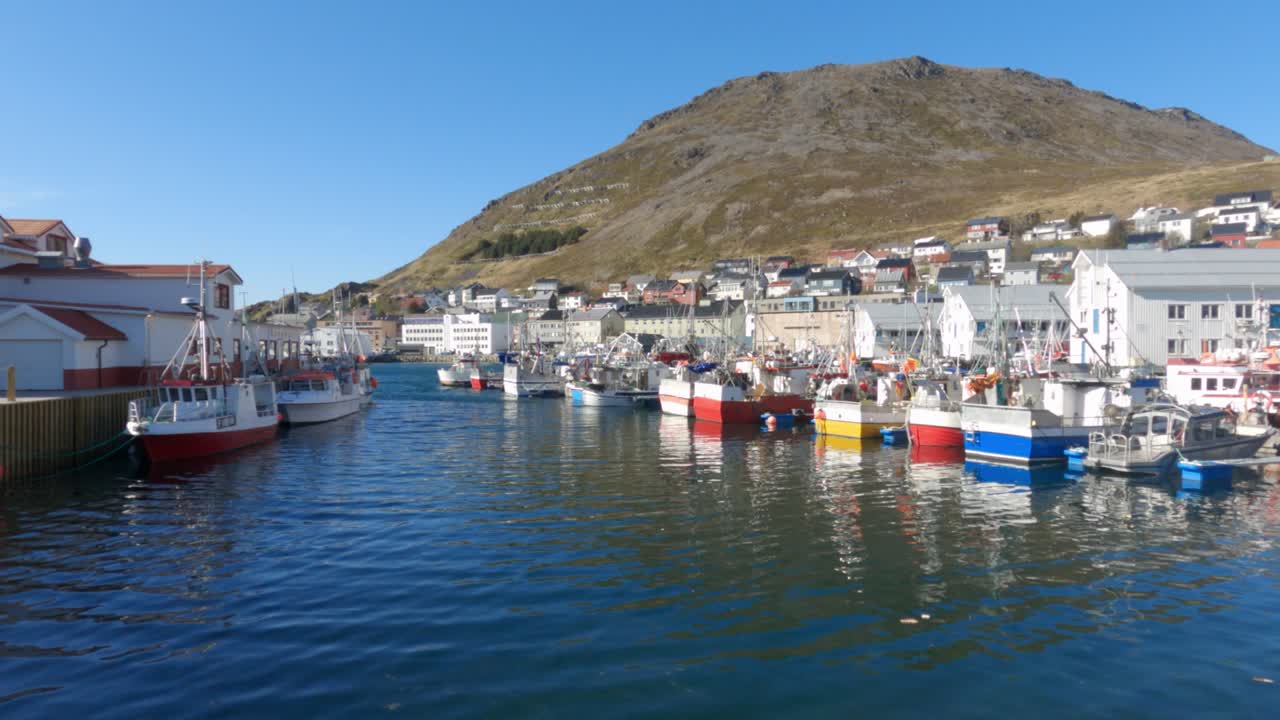 Scenografico porto artico norvegese a Honningsvåg vicino a Capo Nord con colorate barche da pesca ormeggiate lungo la banchina, acqua blu calma e uno sfondo montuoso aspro sotto un cielo estivo sereno