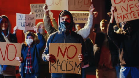 retrato de un hombre caucásico con bufanda en la cara y mirando la cámara mientras sostiene un cartel de "no racismo" en una protesta con un grupo multiétnico de personas en la calle