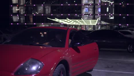 Man-getting-into-red-sport-car-on-drivers-side.-City-lights-at-night-on-the-background