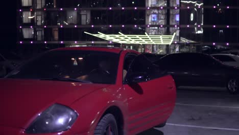 man getting into red sport car on drivers side. city lights at night on the background