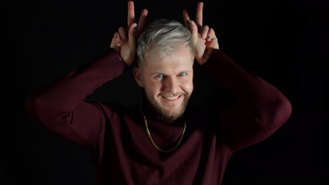 funny joyful man in stylish blouse making bunny ears hand gesture over head on black background