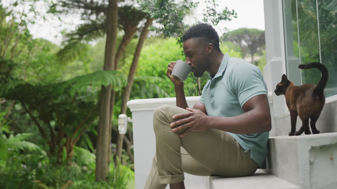 счастливый афроамериканский мужчина сидит с кошкой и пьет кофе в саду